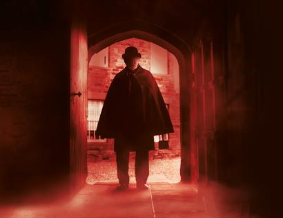 a dark photograph with a near silhouette of a man in a bowler hat and cloak as he enters through an arched wooden door. There is a red hue and a blur.