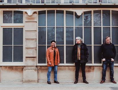 A photograph of three men stood outdoors in front of paned windows.