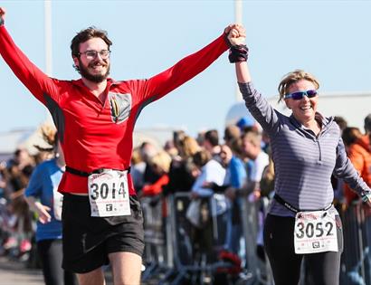 Two runners holding hands cross the finish at the Hastings Half Marathon, wearing race bibs as spectators line the course.