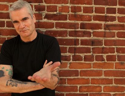 Photograph of white man in black t-shirt with tattoo arms standing against a brick wall.