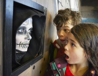 Two shocked looking children to the right look at wall on the left. Wall has a black frame with a human face painted like a skeleton.