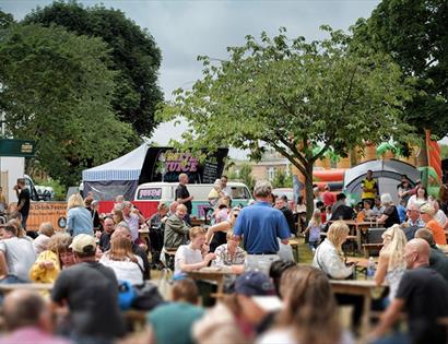 a landscape view of a country festivals. Crowds of people on picnic benches with food bars and a tent in the background.