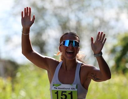 Runner wearing a numbered bib raises both hands while finishing a 10k race outdoors on a sunny day.