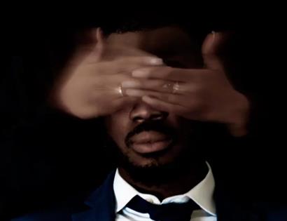 portrait against a black background of black man with a woman's hands covering his eyes.