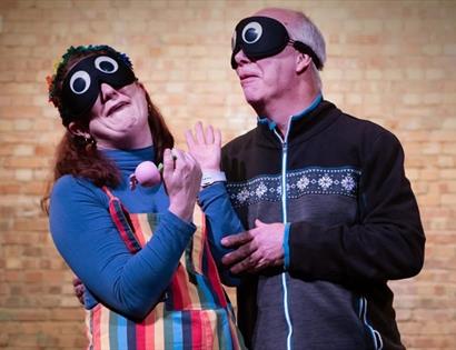 Two performers wearing playful eye masks act together on stage against a brick wall.