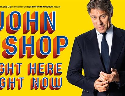 A poster with a yellow background. To the right a white man in a suite. Left says John Bishop. Right here right now.