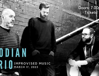 Poster with black and white photograph of three men standing and sitting on steps. Blue text in bottom left hand corner says "Kodian Trio" then "impro