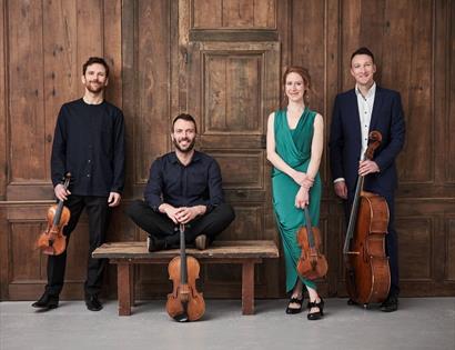 a photograph of four musicians standing in a row against wooden panelled wall. three men in dark suits, one sitting crossed legged on bench, others st