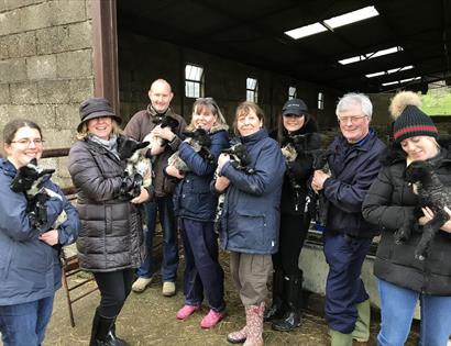 A group of eight adults in coats outside an open barn door, each holding a lamb in their arms.