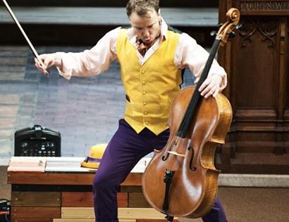 A man in a yellow waistcoat and purple trousers plays a cello on stage. For a Rye Creative Centre event, Rother.
