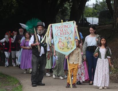 A May Queen procession with a child holding a banner and a chimney sweep at the front.