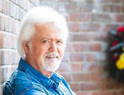 a photograph of Merrill Osmond against a brick wall, flowers in background. A man with white hair and beard.