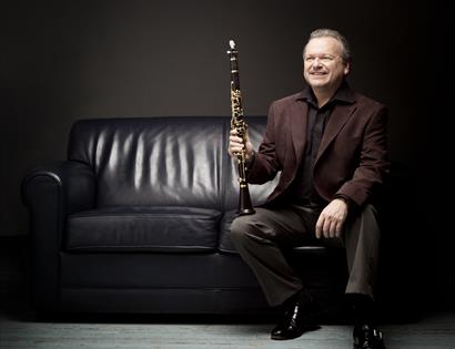 A photograph of a man sat on a black sofa with a clarinet resting on knee. Black background, man wears black suit.
