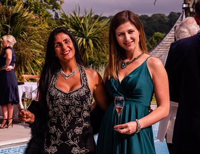 two women arm in arm smiling at the camera and holding champagne flutes. They are wearing dresses and in the background are other smartly dressed peop