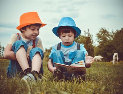 a photograph of two children sat on grass, one wearing an orange trilby hat, the other blue. Boy on right reads a book