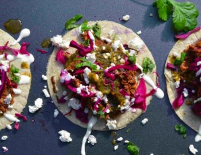 A photograph of three open tacos in a rowwith messy sour cream and coriander leaves over the grey surface. For Taco night at the hub bodiam rother