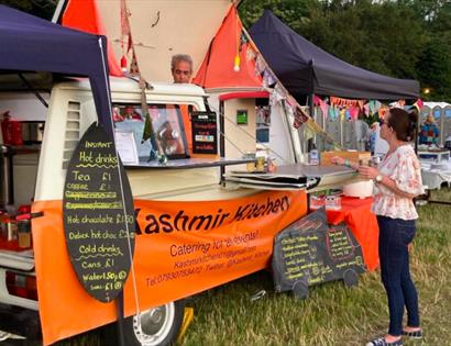 a photograph of a food van in a field with lady standing to make her order. Van has orange banner saying Kashmir Kitchen.