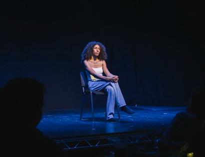 Speaker seated on a chair under blue stage lighting at “Trans Art and Activism: How do we shape the world?” event.