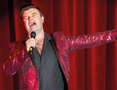 a man singing wearing a shiny red jacket against a red curtain.