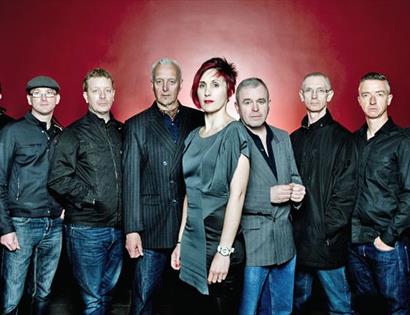 a photograph of the band, the south. nine band members are all lined up before a plain red background. wearing dark suits and coats.
