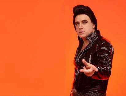 man standing before plain bright orange background. white man in leather jacket and high brown hair, like Elvis Presley.