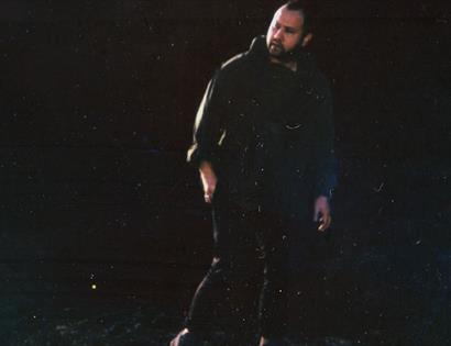 photograph of a white man in a black hoodie, in the shadows against a black background.