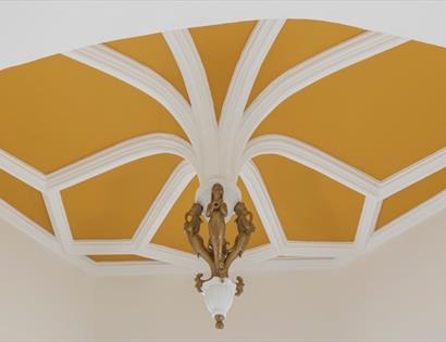 Ornate ceiling with white geometric beams and a gold background, featuring a decorative hanging light.