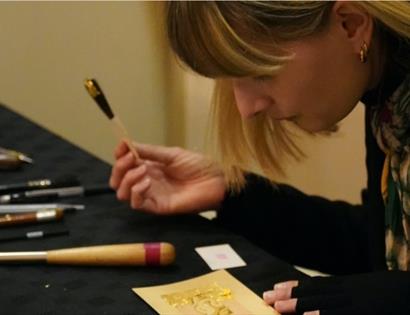 Person working at a table, applying gold leaf to a decorative design using fine tools and brushes.