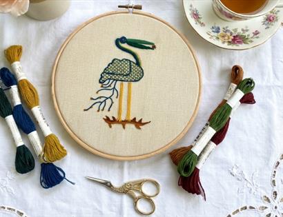 Embroidery hoop with a stitched bird design surrounded by colourful threads, scissors, and a teacup on a lace cloth.