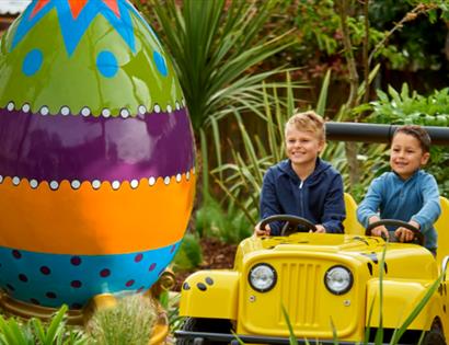 Two children in a yellow jeep ride next to a large colourful egg at Drusillas Park.