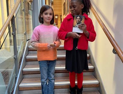 Two girls stand on steps indoors, one clutches a book.