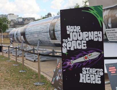 picture of a large replica rocket mounted for display outdoors. A display panel in the foreground says 'Your journey to space starts here'.