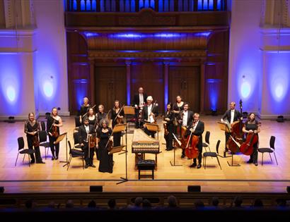 a high up view of an orchestra performing on stage. light wooden flooring, musicians all stand and face toward hidden audience.