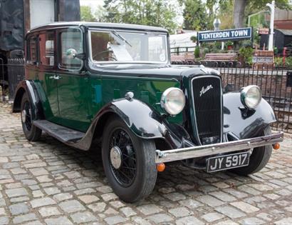 a photograph of a vintage green car parked on brick paving outside a railway station.