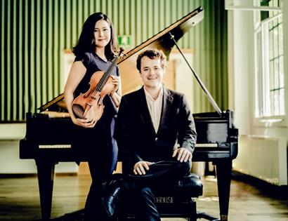 photograph of two musicians, a woman standing with violin and man seated with back against piano, green wallpaper in the background and light coming t