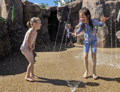Children playing and running through water jets at Knockhatch in an outdoor splash area surrounded by rocky walls.