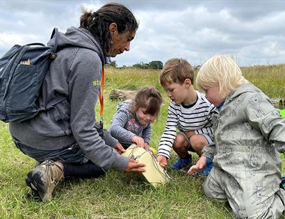 photograph of three small children playing in the grass, woman with backpack showing them a box.