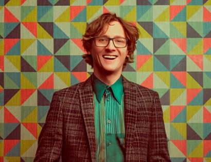A photograph of comedian Ed Byrne against a coloured geometric wallpaper.