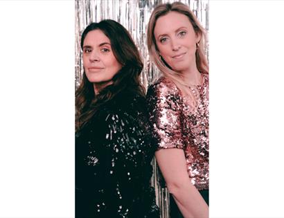 Two people wearing sparkly black and rose-gold sequin tops standing in front of a silver tinsel backdrop.