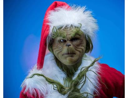 Image shows a picture of 'The Grinch' with green skin wearing a santa outfit which is red with white detail. The background of the image is blue.