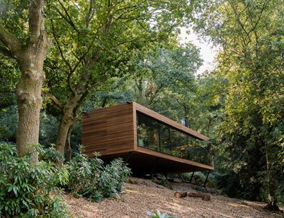 A suspended rectangular wooden lodge with large glass facade in a wooded landscape.