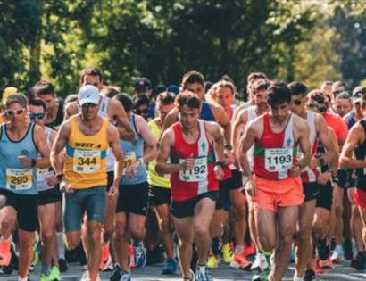Large group of runners in athletic gear starting a race on a sunny outdoor road lined with trees.