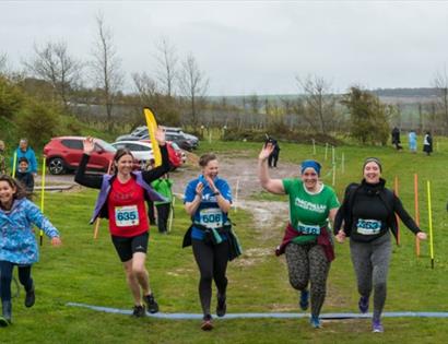 Five runners crossing a finish line on a grassy outdoor course with spectators and cars in the background.