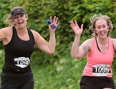 Two runners with race bibs raising their arms while running outdoors on a green, leafy path.
