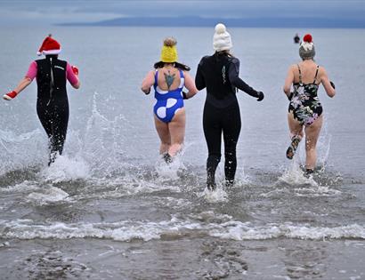 Four people are running into the sea from a sandy shoreline, creating splashes as they enter the water. They are dressed in festive and winter-themed