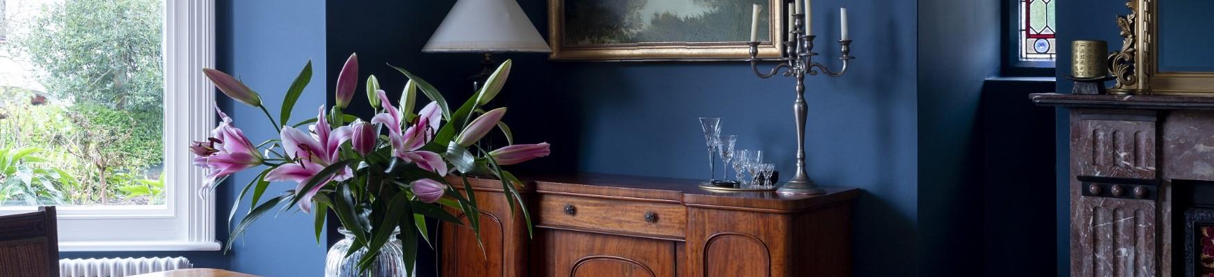 A vintage dining room with deep blue walls, a wooden table, and a vase of pink lilies, with natural light coming in from a large window in The Knowle Hotel in St Leonards.
