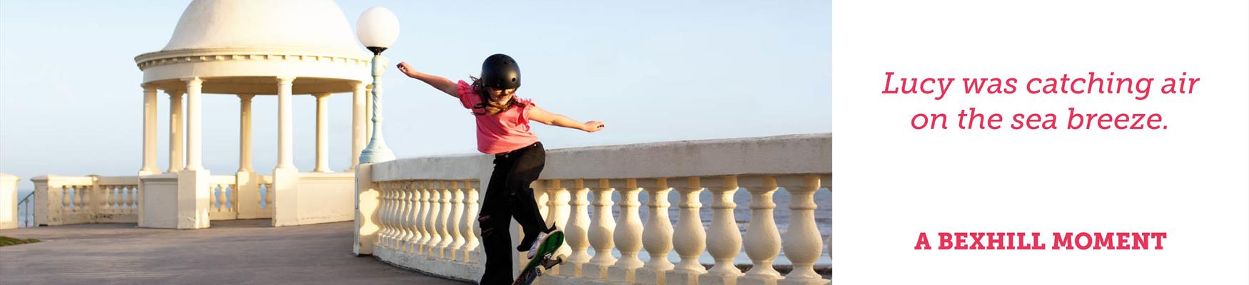 A photograph of a young girl skating at Bexhill on Sea seafront on a sunny day. She wears a helmet and her arms are outstretched. She is on a concrete terrace with a domed white structure in the background. The caption next to the photograph reads “Lucy was catching the air on the sea breeze”.
