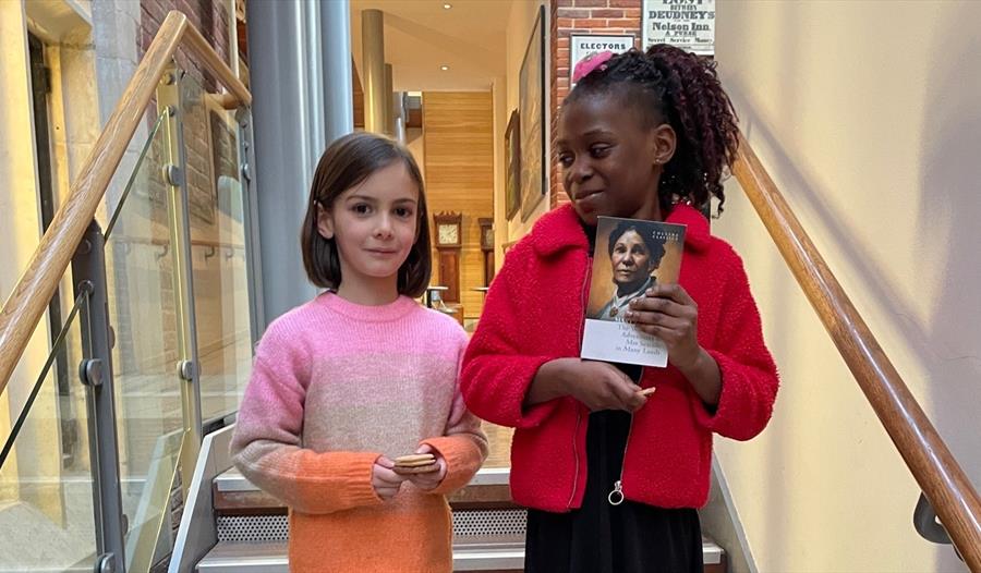 Two girls stand on steps indoors, one clutches a book.