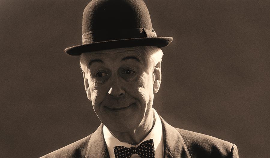 a black and white photograph of a white man, showing his head only. He wears a bowler hat, bow tie and has painted on eyebrows.