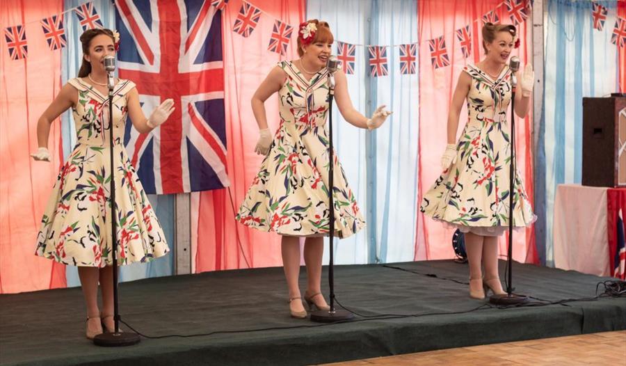 three women on stage in vintage swing dressers singing. Hair in 1940s style. Union jack flag and bunting in background.